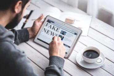 Un homme sur une tablette lit de l'information. Le contenu de l'écran indique fake news. Une tasse de café est posée à côté.