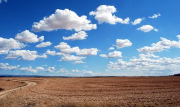 Un grand champ avec une route de campagne sur la gauche et un ciel bleu avec des nuages à perte de vue.