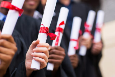 Une rangée d'étudiants tiennent leur diplôme d'une main, enroulée comme un parchemin avec un ruban rouge pour le retenir.