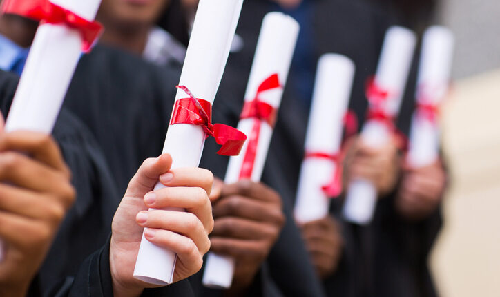 Une rangée d'étudiants tiennent leur diplôme d'une main, enroulée comme un parchemin avec un ruban rouge pour le retenir.